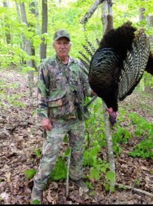 "A youthful Roscoe Spaulding in hunting attire, holding a turkey call in a forest setting."