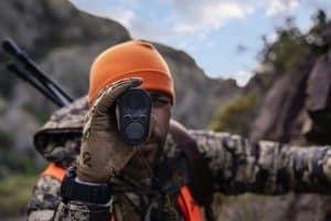 Hunter in camouflage with orange beanie aims a handheld rangefinder at the camera, outdoors among rocks and trees (informative).