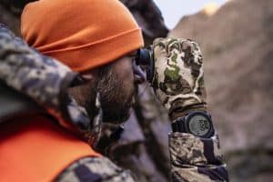 Man in camouflage gear and an orange beanie looks through a monocular or small scope, outdoors in a rocky area.