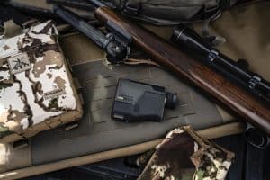Rifle with scope and wooden stock resting on camouflage gear on a padded surface; a Garmin handheld device sits nearby on the mat.
