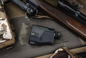 Field Tested: Garmin Xero L60i Rangefinder Review Rifle with scope and wooden stock resting on camouflage gear on a padded surface; a Garmin handheld device sits nearby on the mat.