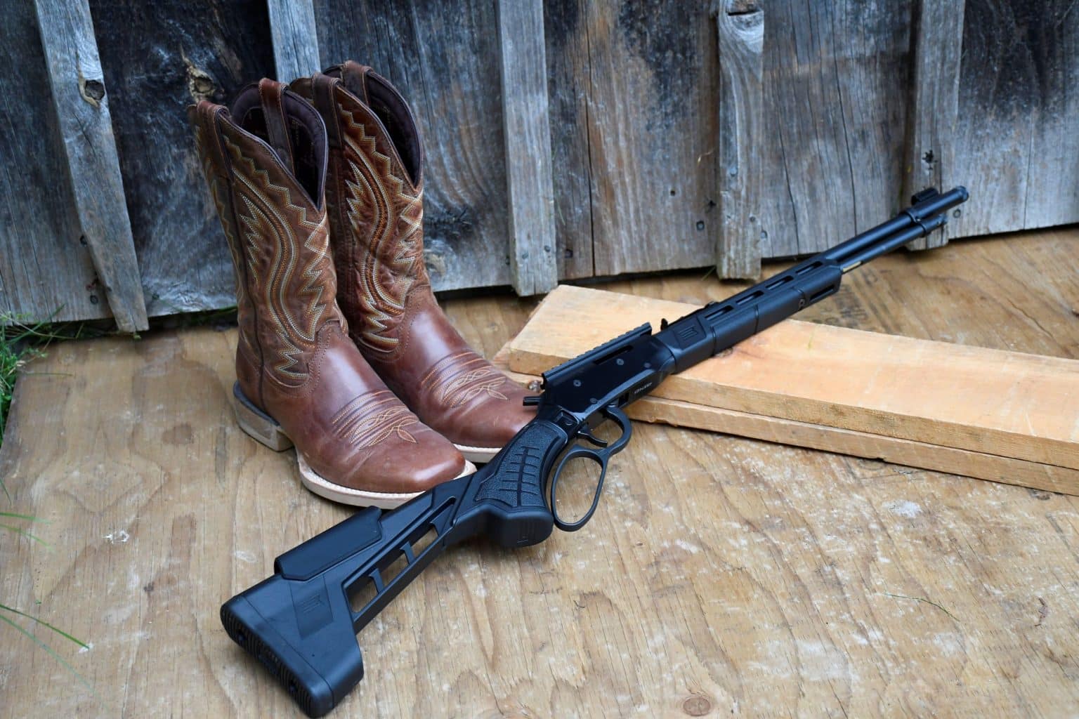 Savage Revel Tactical lever-action rimfire rifle leaning against a rustic wooden wall beside cowboy boots.