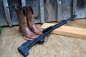 Savage Revel Tactical lever-action rimfire rifle leaning against a rustic wooden wall beside cowboy boots.
